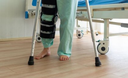 The legs of a person by a hospital bed in green clothing with a braced knee and crutches